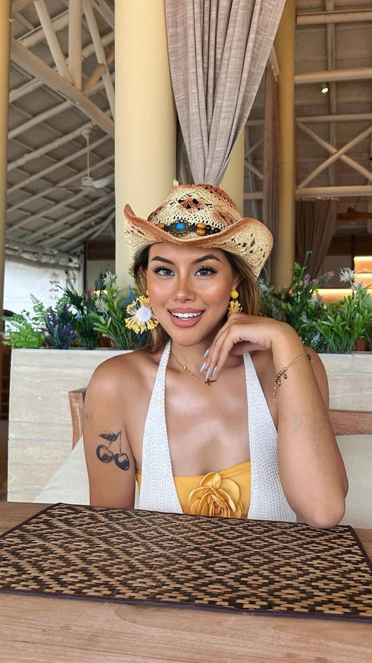 Woman with a radiant smile wearing a cowboy hat and floral earrings, showcasing a bold temporary cherry tattoo on her upper arm in a tropical indoor setting.