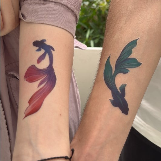 A close-up of two people's arms, showing matching tattoos of a red-purple fish and a green-blue fish.