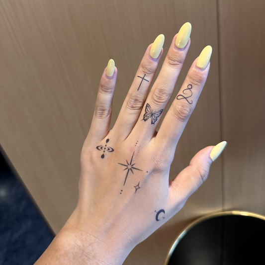 A close-up of a hand with yellow almond-shaped nails, featuring several minimalist tattoos