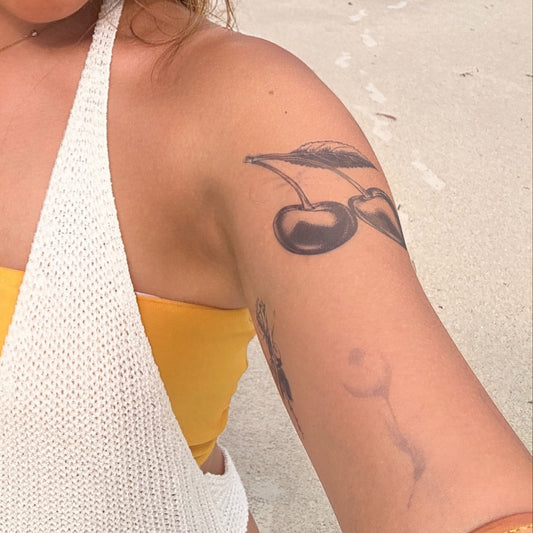 SteezyInk Twin Cherries Temporary Tattoo on arm, beach background, white top. Represents sweetness, love, and vitality.